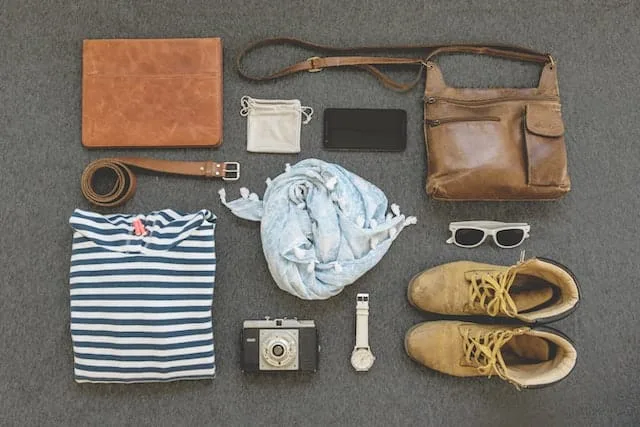 A flat lay of travel essentials on a gray background: leather accessories, striped shirt, light scarf, boots, vintage camera, watch, sunglasses, and small pouches. The arrangement showcases a curated collection for a stylish traveler, blending functionality with fashion.