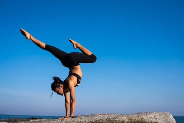 A woman performs a handstand on a rocky surface by the ocean. She is wearing black leggings and a black sports bra. Her legs are split in a V-shape against a clear blue sky. The pose demonstrates strength, balance, and flexibility in an outdoor yoga or acrobatic practice.