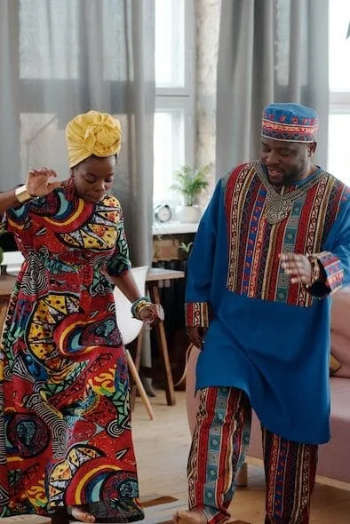 A man and woman joyfully dancing together indoors. The woman wears a colorful, patterned dress and a yellow headwrap, while the man wears a traditional blue outfit with vibrant, intricate designs. They are in a bright room with large windows and casual furniture.