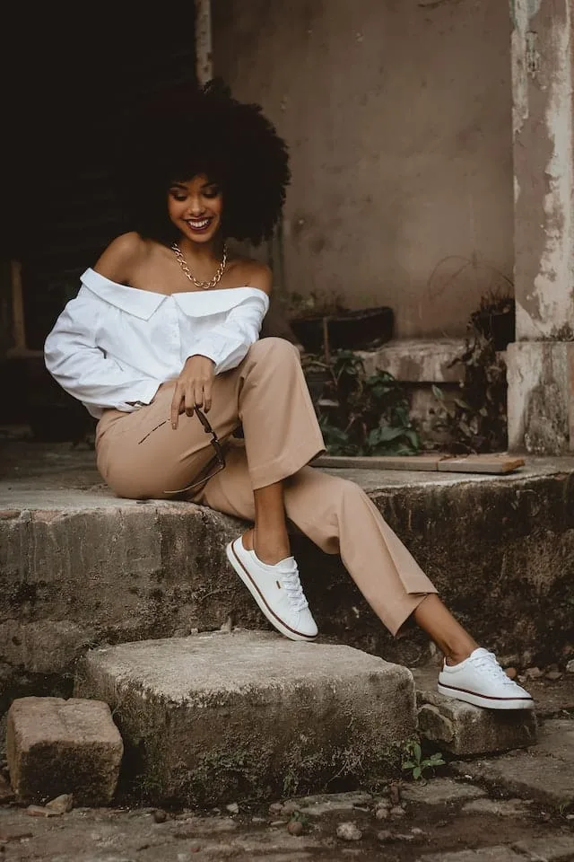 A stylish woman with natural curly hair sits casually on an outdoor step. She wears a white off-shoulder blouse, beige trousers, and white sneakers, accessorized with a gold chain necklace. The background features a rustic, worn wall and plants growing around the steps.