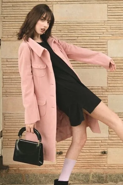A young woman in a dynamic pose, wearing a long pale pink coat over a black dress, accessorized with a small black handbag. She stands against a textured stone wall, with one leg extended in a playful stance."