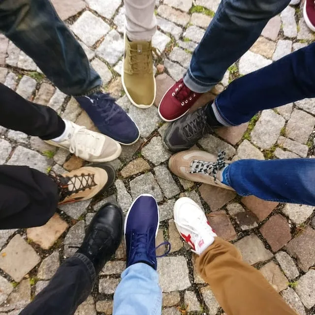 The image shows a group of people standing in a circle, with their feet pointed toward the center. Each person is wearing a different style of sneakers, ranging from casual to sporty designs, in various colors such as blue, black, beige, red, and white. The ground beneath them is made of cobblestone, adding a rustic feel to the scene. The shoes reflect diversity in style and personality.