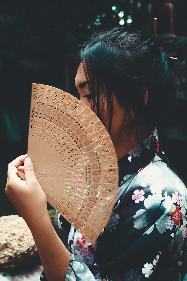 Close-up of a woman in a floral kimono holding an ornate wooden folding fan in front of her face. Her dark hair is styled in a traditional updo with decorative pins. The intricate patterns of the fan partially obscure her features, creating an elegant and mysterious atmosphere. The background is dark, emphasizing the subject and the cultural elements of the image.