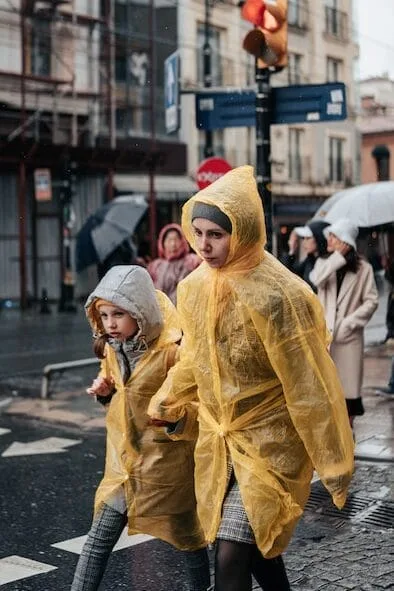 A woman and child cross a wet city street in the rain, wearing bright yellow plastic ponchos. The woman holds the child's hand as they walk. Behind them, other pedestrians with umbrellas can be seen. The scene captures a rainy day in an urban setting with traffic lights and buildings visible in the background.