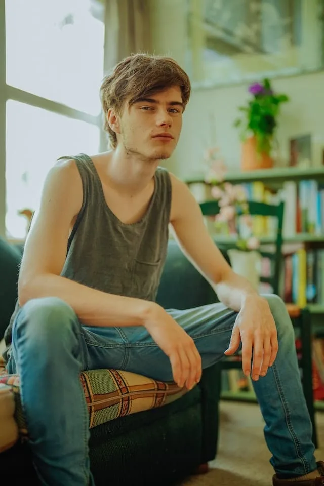 A young man with tousled brown hair sits casually in an armchair, wearing a gray tank top and blue jeans. He's in a cozy home setting with a bookshelf and potted plant visible in the background. The soft lighting gives the image a warm, intimate atmosphere.