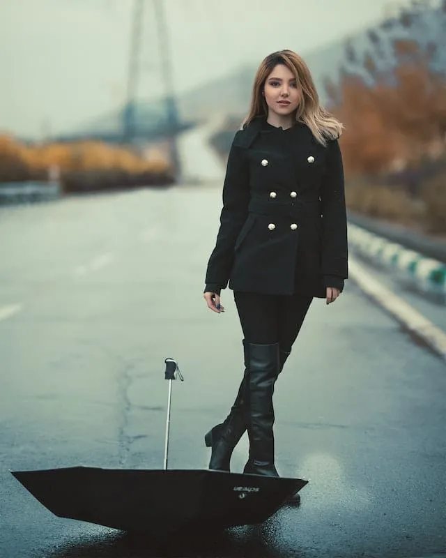A young woman stands on a wet street near a body of water. She wears a black double-breasted coat, dark pants, and knee-high boots. Her blonde hair is styled in loose waves. In the foreground, an upturned black umbrella sits on the wet pavement. The background shows blurred autumn foliage and a bridge or structure in the distance.