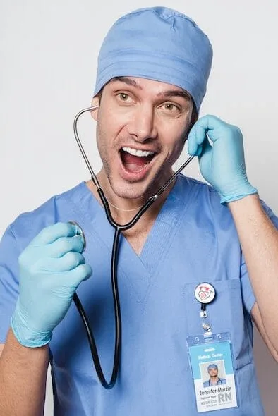 The image shows a male nurse in blue scrubs, wearing a blue surgical cap and gloves. He is playfully holding a stethoscope to his mouth as if he's about to speak into it, with an enthusiastic and humorous expression. He has a name badge identifying him as "Jennifer Martin" and a pin on his scrubs. The background is plain white, emphasizing the light-hearted, fun pose.