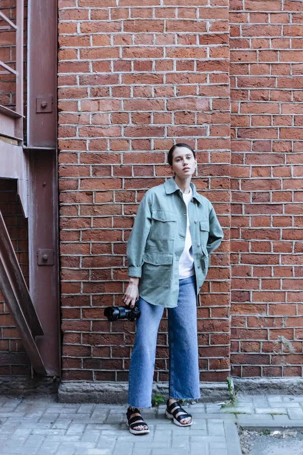 A young woman stands against a brick wall, wearing a sage green oversized jacket, white shirt, and blue jeans. She holds a camera and wears sandals. The urban setting features a brown drainpipe and paved sidewalk, creating a casual street photography aesthetic.