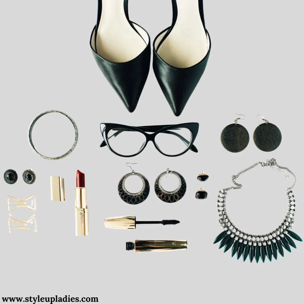 This image displays a carefully curated selection of women's fashion accessories on a light gray background. Featured items include a pair of classic black pointed heels, large black-framed glasses, and a variety of jewelry such as a simple silver bracelet, two pairs of earrings—one round and black, the other silver and hoop-styled—, and a striking necklace with a black and silver design. The arrangement also includes makeup essentials: a red lipstick, a gold lipstick tube, and black mascara. These accessories are essential for completing a chic and sophisticated formal look.